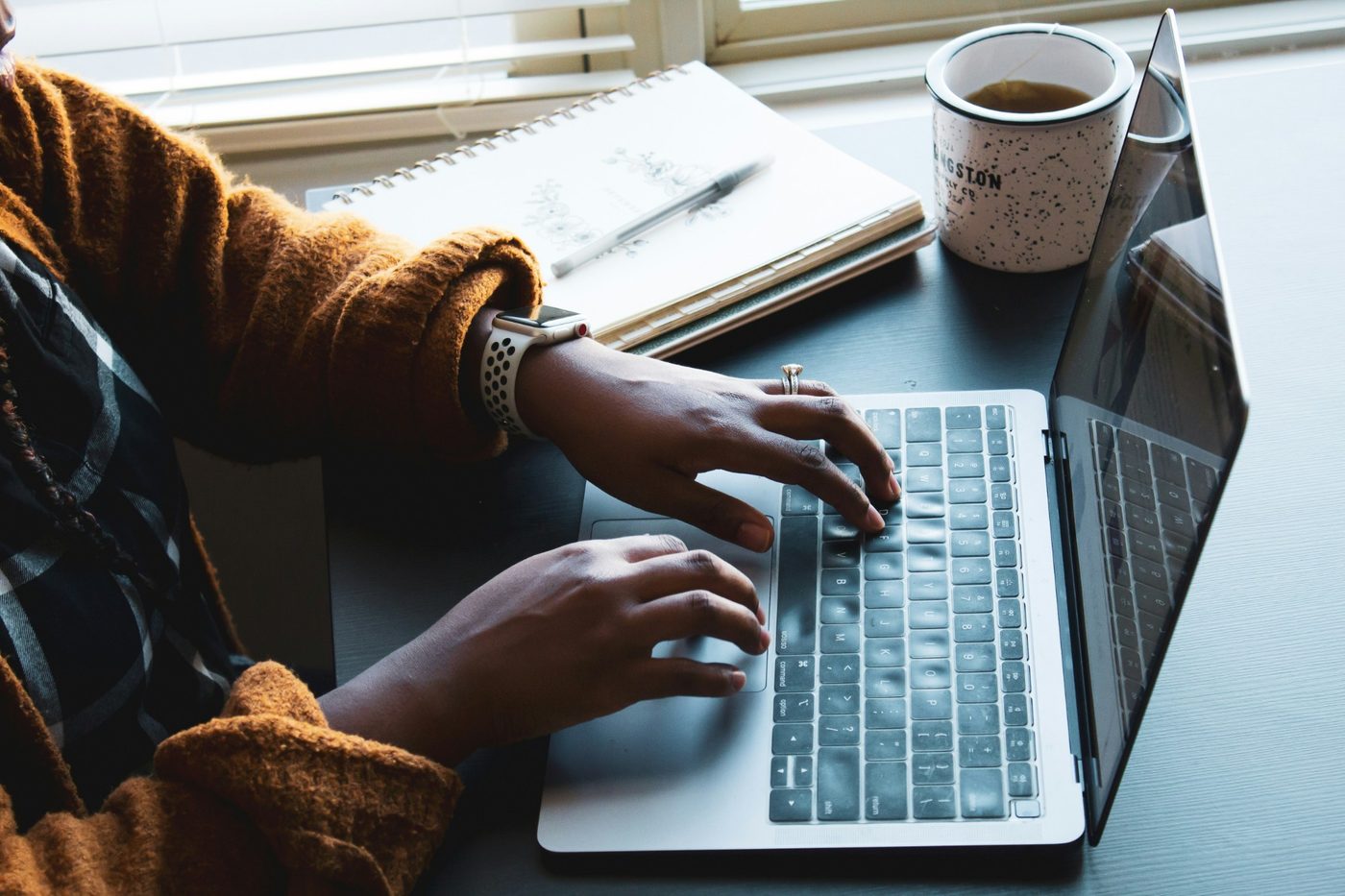 Hands typing on a laptop beside a notebook and a cup of tea